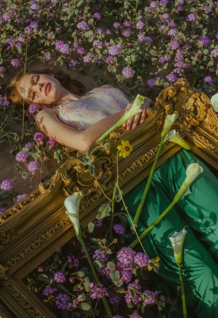 Woman laying on an ornate gold frame in a field of small purple wildflowers. She is surrounded by white calla lily flowers.