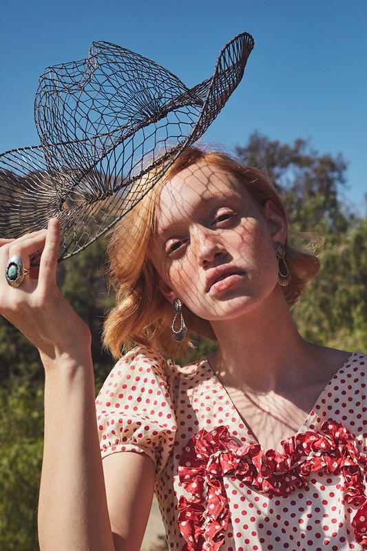 A young woman in a red-and-white polka dot dress poses outdoors with a black wire hat, under the bright daylight, surrounded by nature.
