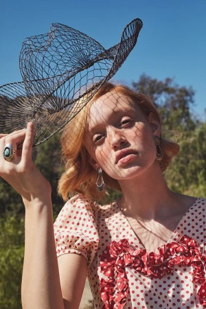 A young woman in a red-and-white polka dot dress poses outdoors with a black wire hat, under the bright daylight, surrounded by nature.