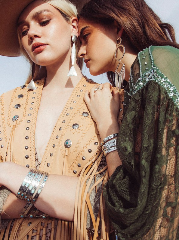 Two fashion models in Western attire featuring tan vest with fringe and green sheer lace top with silver bracelets and earrings, outdoors.