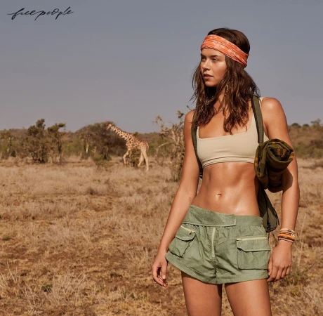 Adult woman in athleisure apparel poses in a savanna environment with a giraffe in the background, outdoors on a sunny day.