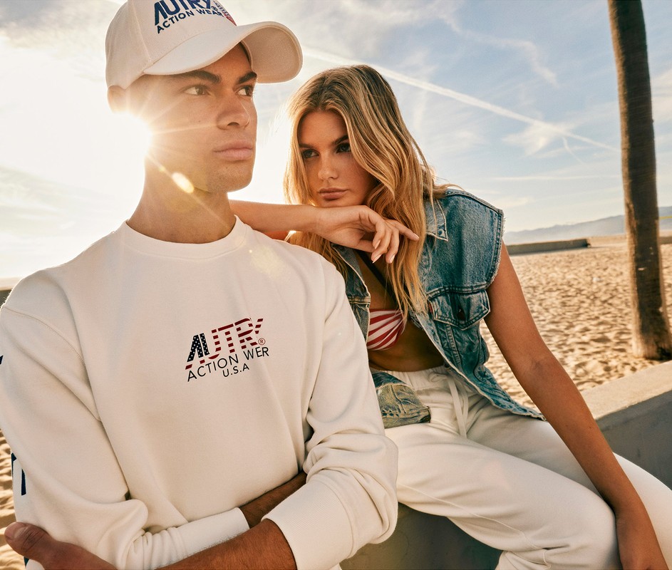 Two fashion models on a beach, wearing Autry Action Wear branded clothing and posing under a palm tree and blue sky in the sunshine.
