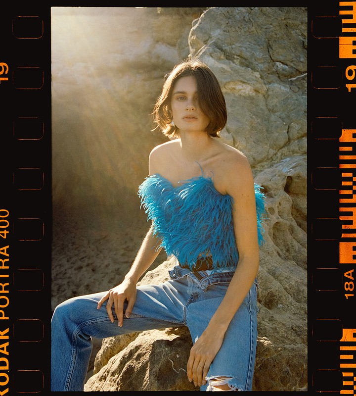 A woman in a blue feathered top and jeans is sitting on a rock, with sunlight and a film border surrounding her. Fashion shot outdoors.