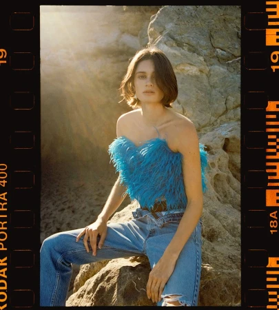 A woman in a blue feathered top and jeans is sitting on a rock, with sunlight and a film border surrounding her. Fashion shot outdoors.