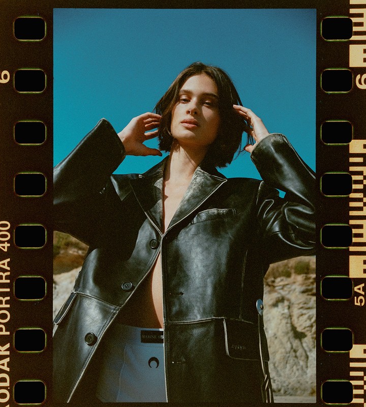 A fashionable adult woman poses outdoors with a black leather jacket against a blue sky and sandy background, captured on film stock.
