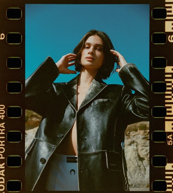 A fashionable adult woman poses outdoors with a black leather jacket against a blue sky and sandy background, captured on film stock.