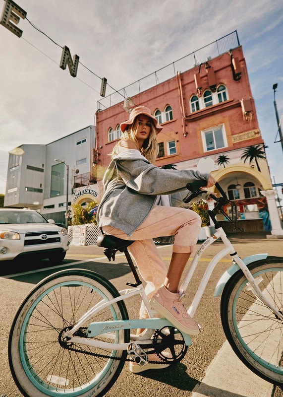 A blonde woman rides a bicycle on a city street with peach-colored buildings under a bright blue sky with white clouds during daylight hours.