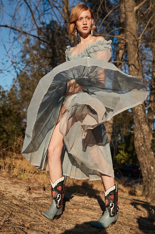 A woman with red hair wears a blue, translucent dress and cowboy boots outdoors, with trees and sky visible in the background.