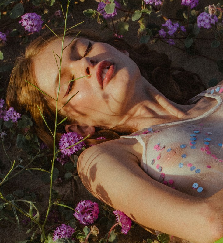 Close-up portrait of a woman lying among purple wildflowers with sparkling sequins on her skin.