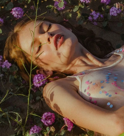 Close-up portrait of a woman lying among purple wildflowers with sparkling sequins on her skin.