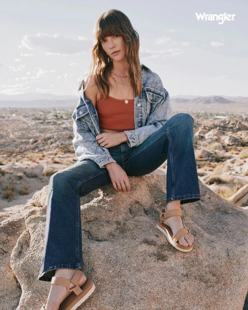 A woman poses in Wrangler jeans and a denim jacket on desert rocks, showcasing a lifestyle fashion image with a bright, natural setting.