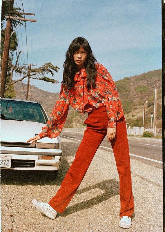 A woman in a floral blouse and red pants poses next to a white car on a roadside with mountains in the background under a clear sky.