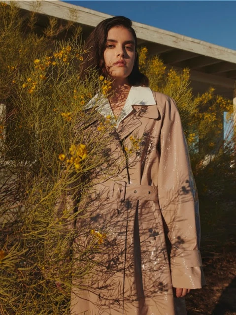 Brunette woman in a tan trench coat stands near a desert plant with yellow flowers on a sunny day outdoors in an editorial fashion shot.
