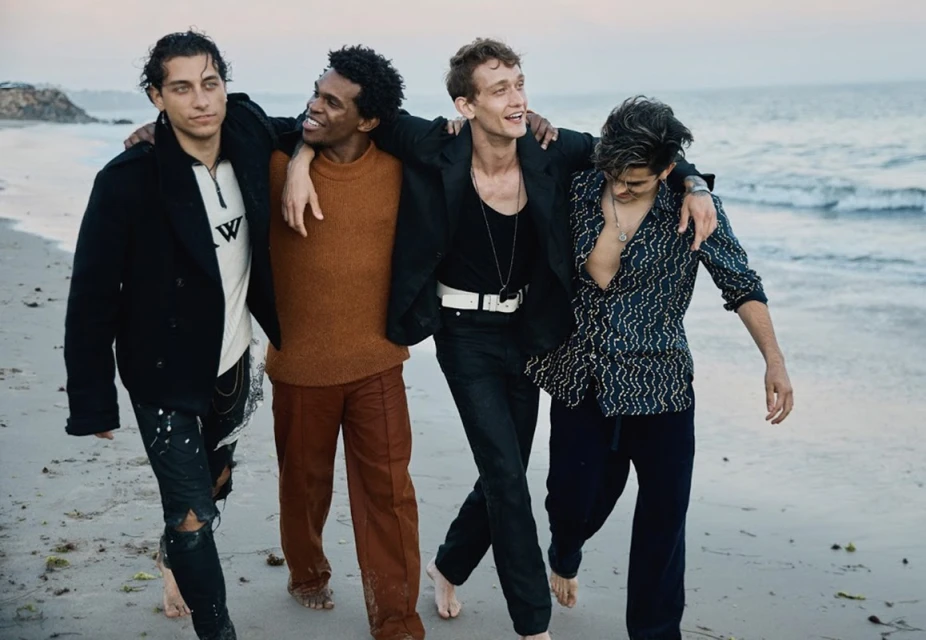 Four adult male models walking together barefoot on a sandy beach near the ocean, wearing stylish fall clothing at daytime.
