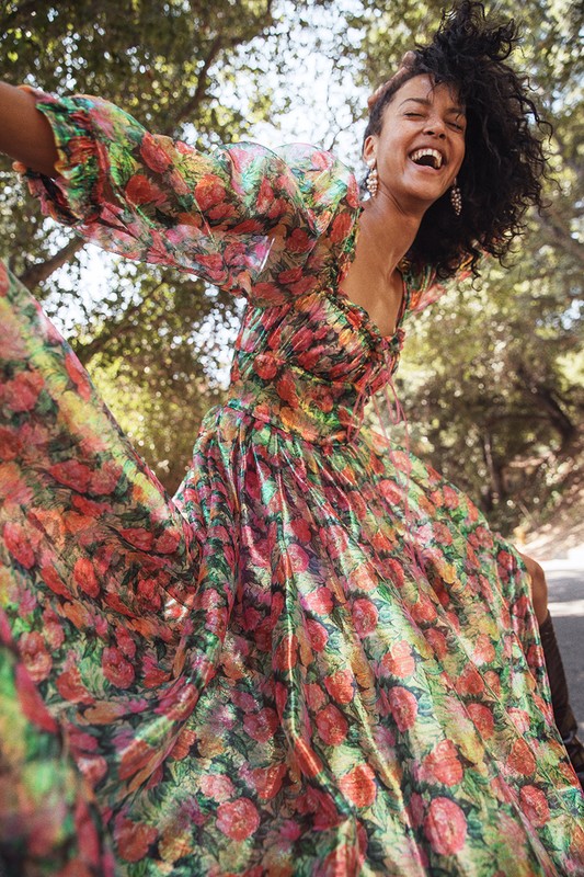 Woman with curly hair in a long floral dress and earrings, outdoors surrounded by trees, smiling and looking up with arms out wide.
