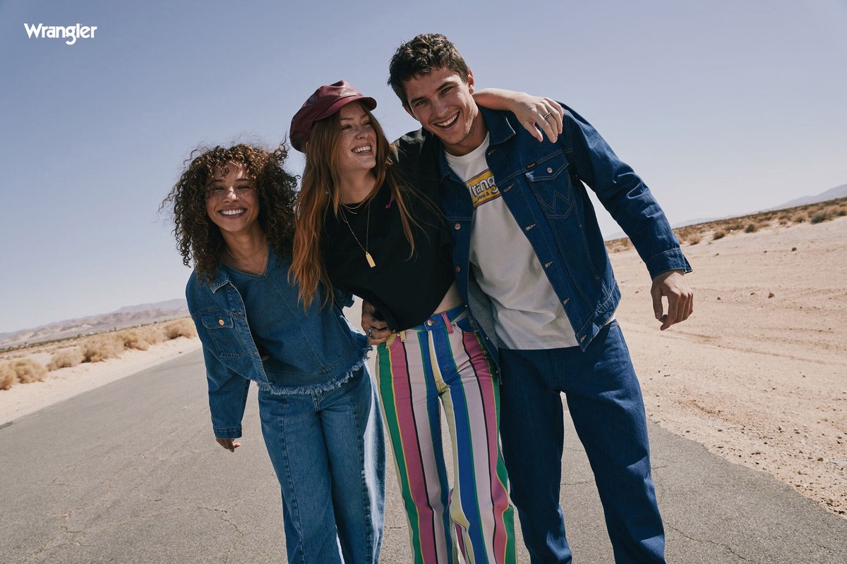 Three smiling models walking together on an open desert road, wearing Wrangler denim jackets and colorful apparel under a clear sky.