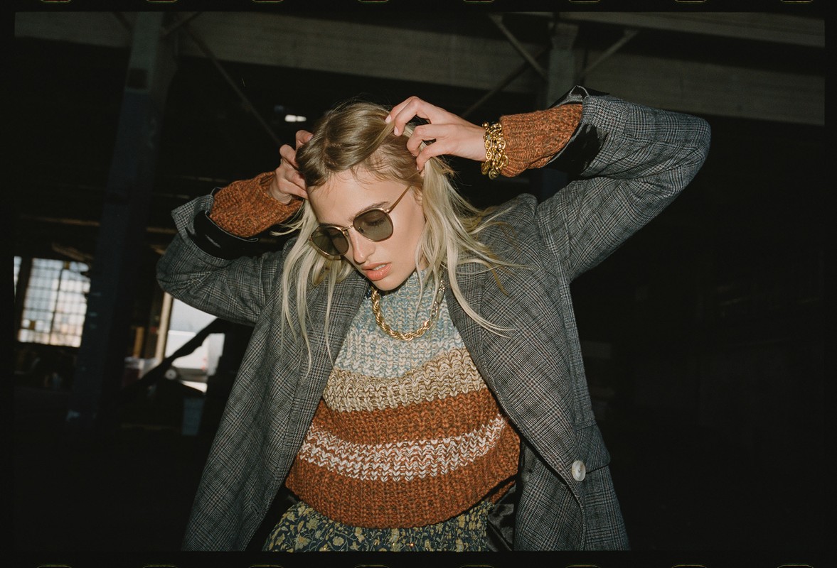 A blonde-haired woman in sunglasses poses in a gray plaid jacket with a colorful knit top, gold necklace, and bracelet in a dark indoor setting.