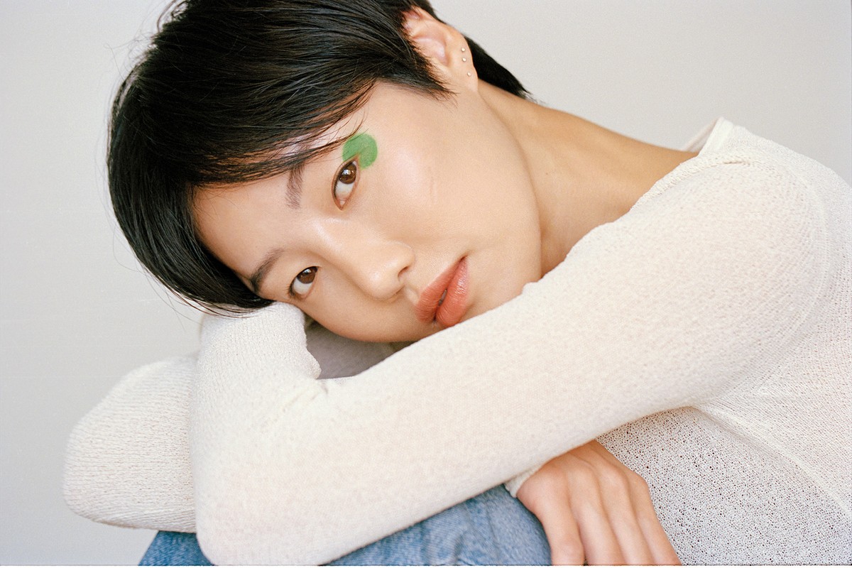 Close-up portrait of a woman with short dark hair wearing a white textured top, with green eye makeup detail.