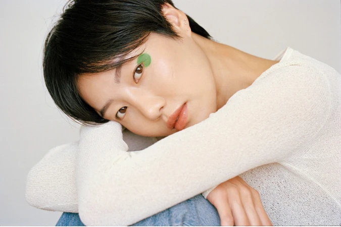 Close-up portrait of a woman with short dark hair wearing a white textured top, with green eye makeup detail.