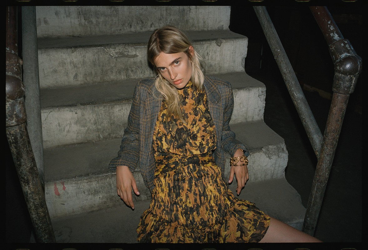 Woman in a patterned dress and blazer poses on concrete stairs. Neutral colors, dark lighting, and a fashion editorial style.