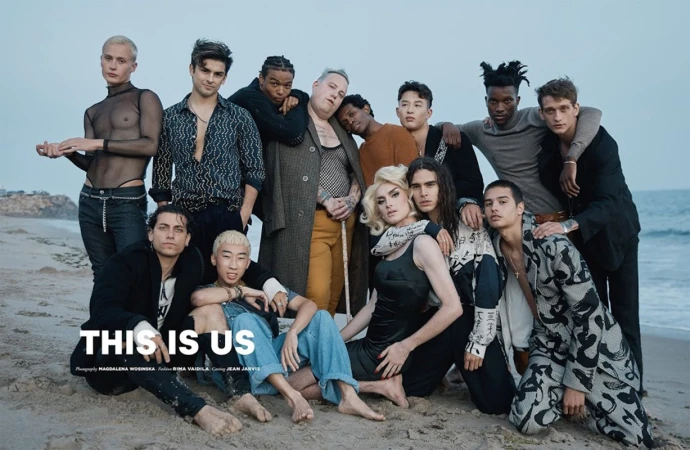 Diverse group of fourteen people poses on a sandy beach with the ocean in the background, creating a cohesive and inclusive portrait.