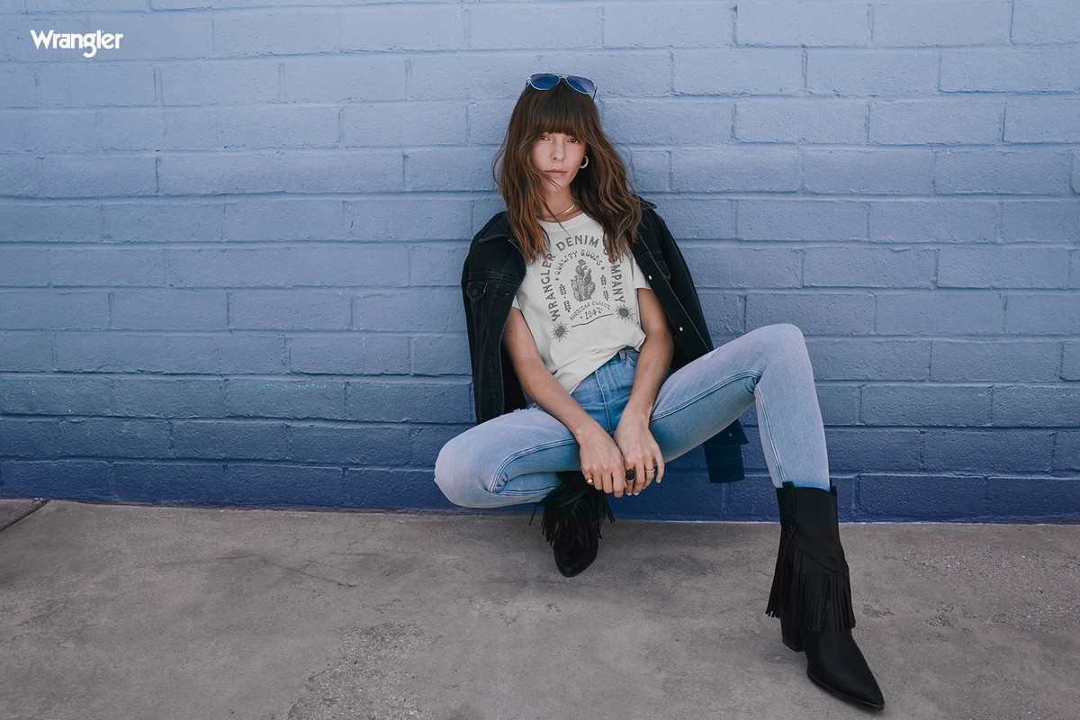 A woman poses in denim jeans, jacket, and fringed boots in front of a blue brick wall, wearing a Wrangler logo t-shirt and sunglasses.