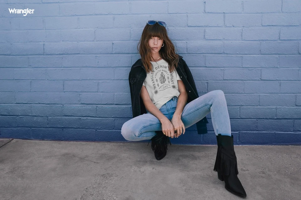 A woman poses in denim jeans, jacket, and fringed boots in front of a blue brick wall, wearing a Wrangler logo t-shirt and sunglasses.