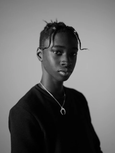 Striking black and white portrait of a young adult with braided hair, wearing a crescent pendant, against a neutral grey backdrop.
