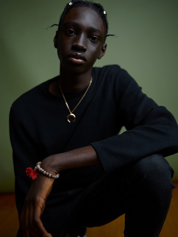 Portrait of a dark-skinned young adult in black clothing, kneeling against a green background, wearing a gold necklace and beaded bracelet.