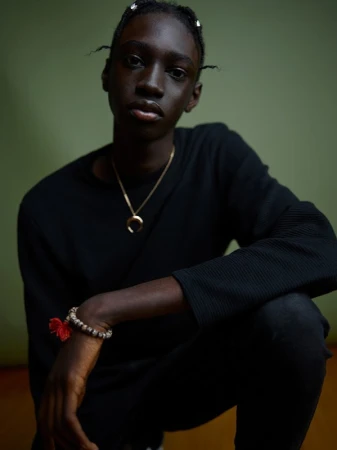 Portrait of a Black model in a black long-sleeve top wearing a gold crescent necklace and beaded bracelet against a dark green backdrop