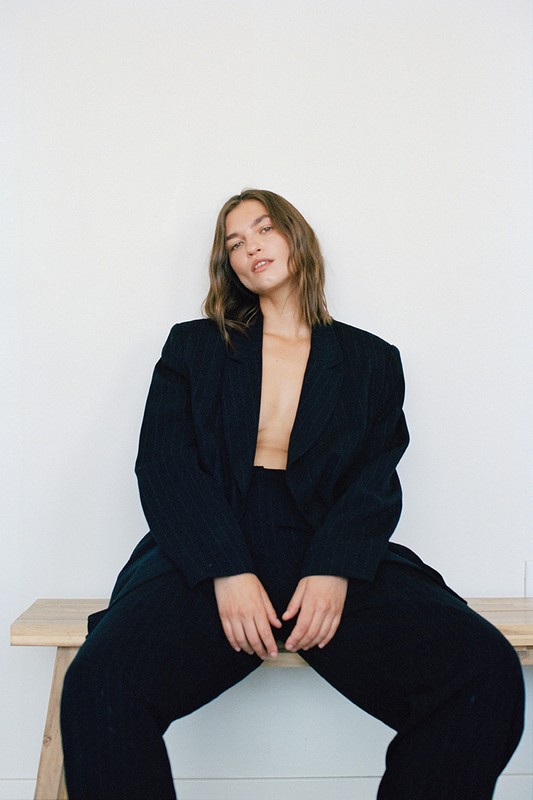 Woman in oversized black suit sits on wooden bench against a white background in a studio setting with natural lighting.