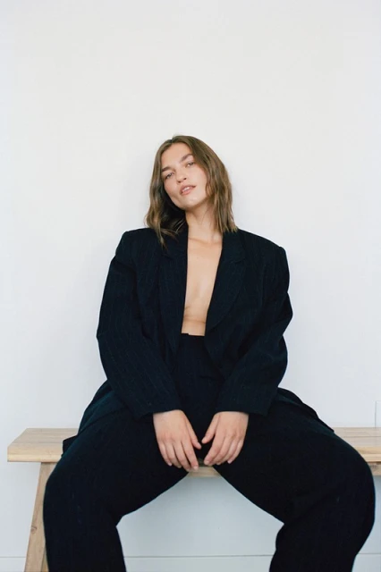 Woman in oversized black suit sits on wooden bench against a white background in a studio setting with natural lighting.