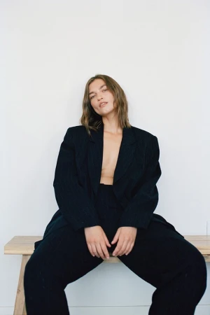 Model with shoulder-length brown hair wearing an oversized black blazer and matching trousers, seated on a wooden bench.