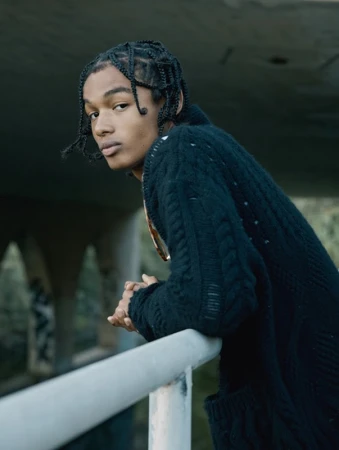Portrait of a young African-American man with braided hair leaning against a gray railing outdoors wearing a black cable knit sweater.