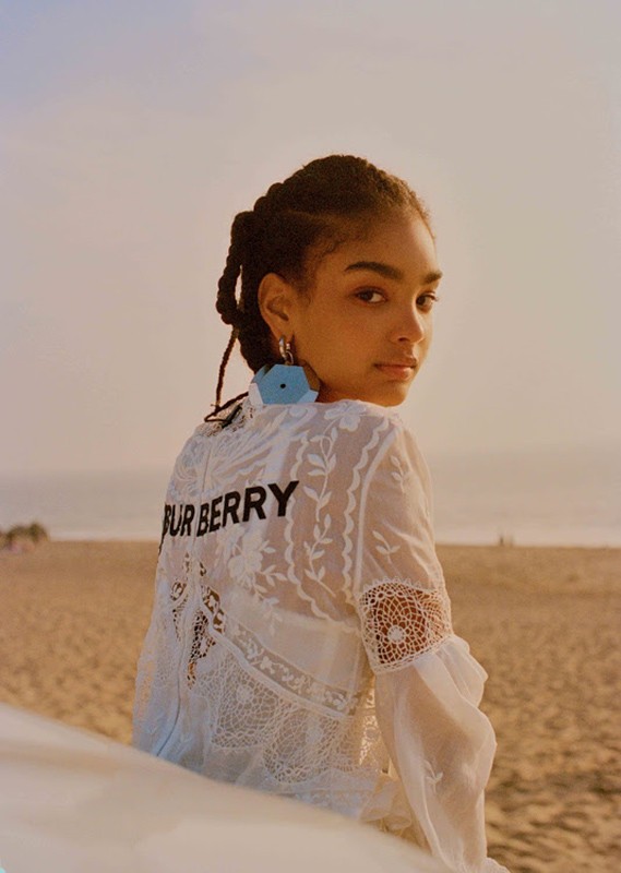 A young woman with braided hair wearing a white lace top with Burberry lettering looks back over her shoulder on a sandy beach at sunset.