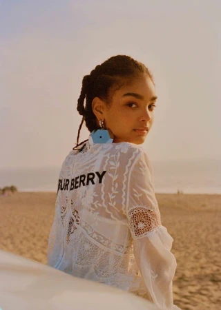 A young woman with braided hair wearing a white lace top with Burberry lettering looks back over her shoulder on a sandy beach at sunset.