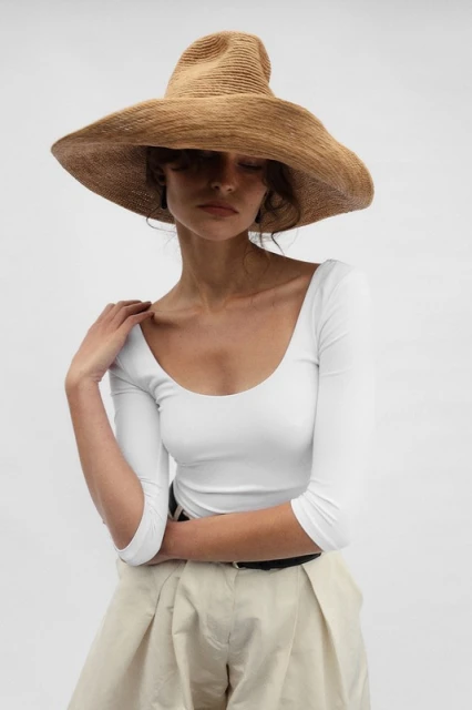 Adult female in a white top and tan pleated shorts poses in a photo studio wearing a wide-brimmed straw sun hat.