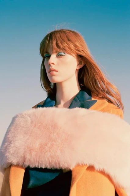 Fashion portrait of a young adult woman with auburn hair and green eyeshadow wearing a yellow jacket with a pink fur stole.
