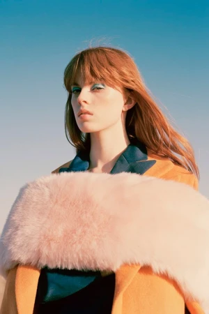 Woman with long red hair wearing an orange coat and pink faux fur stole against a clear blue sky
