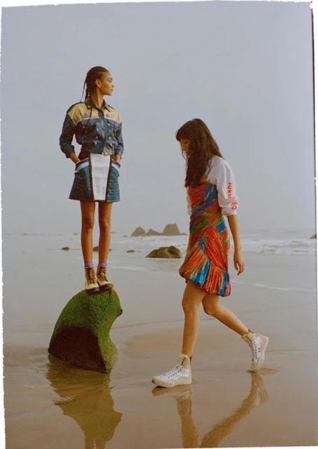 Two women in stylish outfits pose on a beach; one stands on a mossy rock as the other walks through the water.
