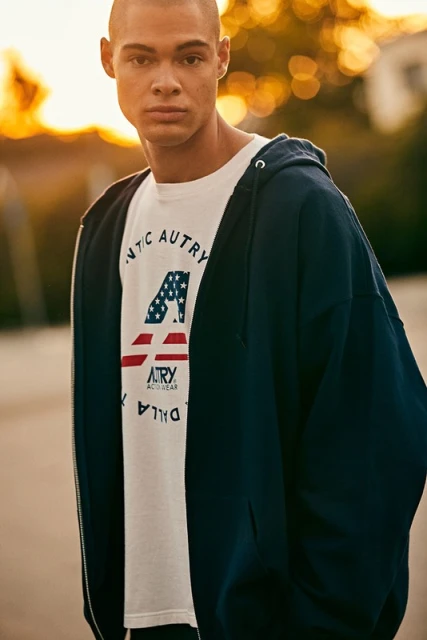 Man in a navy hooded sweatshirt with a white graphic t-shirt, standing outdoors with blurred golden hour background, looking at the camera.