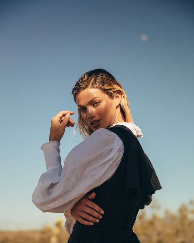 Portrait of a fair-skinned woman in a black vest and white blouse against a blue sky with desert vegetation in the background.
