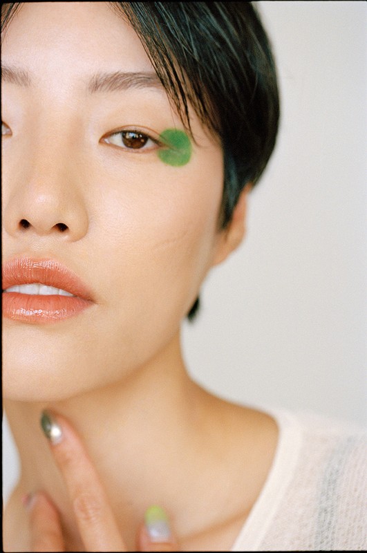Close-up portrait of an Asian woman with short dark hair and a green leaf adhered to her face near her right eye. Neutral background.