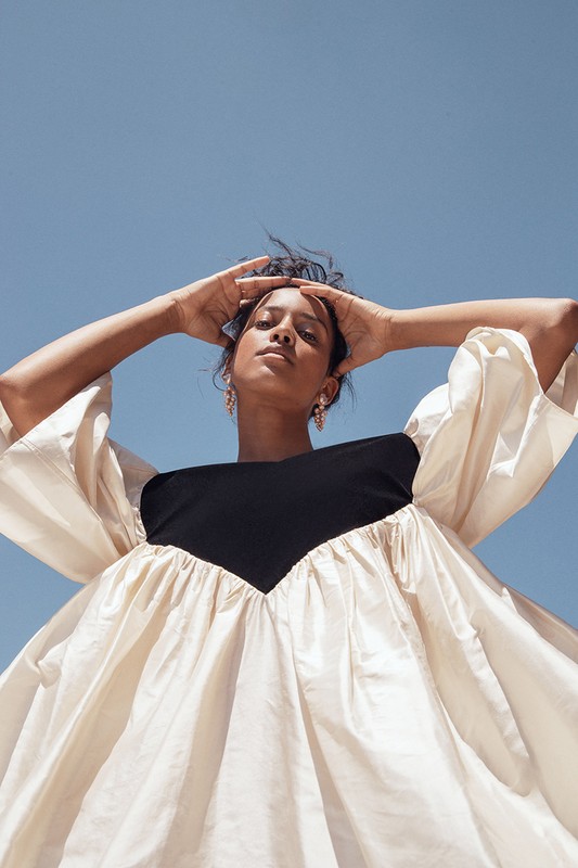 Eye-level shot of adult female model posing outdoors with hands on head and wearing a black and ivory dress against a blue sky.