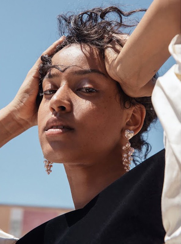Close-up of a young woman with short dark hair and statement earrings posing outdoors against a clear blue sky in natural light.