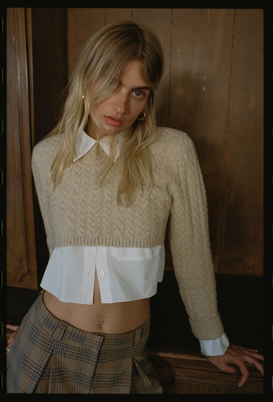 Young woman posing in a beige cropped sweater, white collared shirt and plaid skirt, with wood paneling in the background.