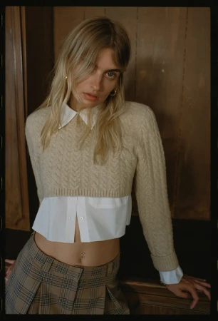 Portrait of a blonde woman wearing a beige cable-knit cropped sweater over a white collared shirt and plaid trousers.