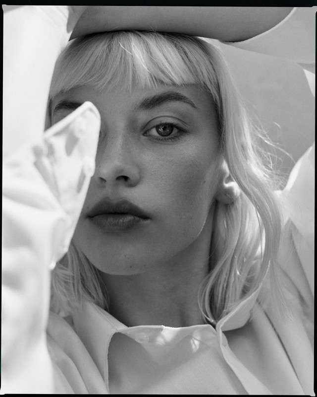 Close-up black and white portrait of a blonde woman with bangs framing her face while holding her arms up, wearing a white shirt.