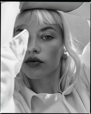Close-up black and white portrait of a blonde woman with bangs framing her face while holding her arms up, wearing a white shirt.