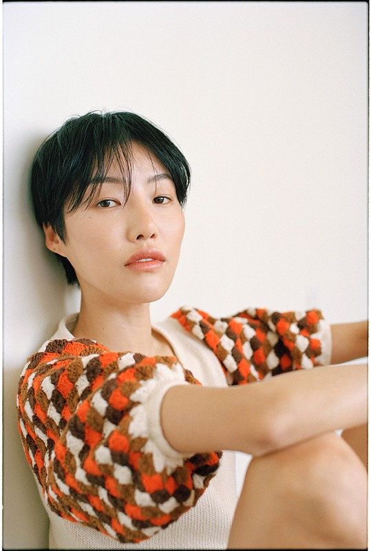 Portrait of an Asian woman with short black hair and wearing a tan and brown checkered top, sitting in front of a plain white wall.